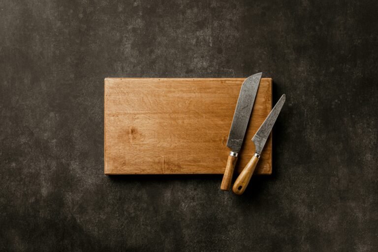 A solid wooden cutting board with two vintage chef knives on a dark rustic surface